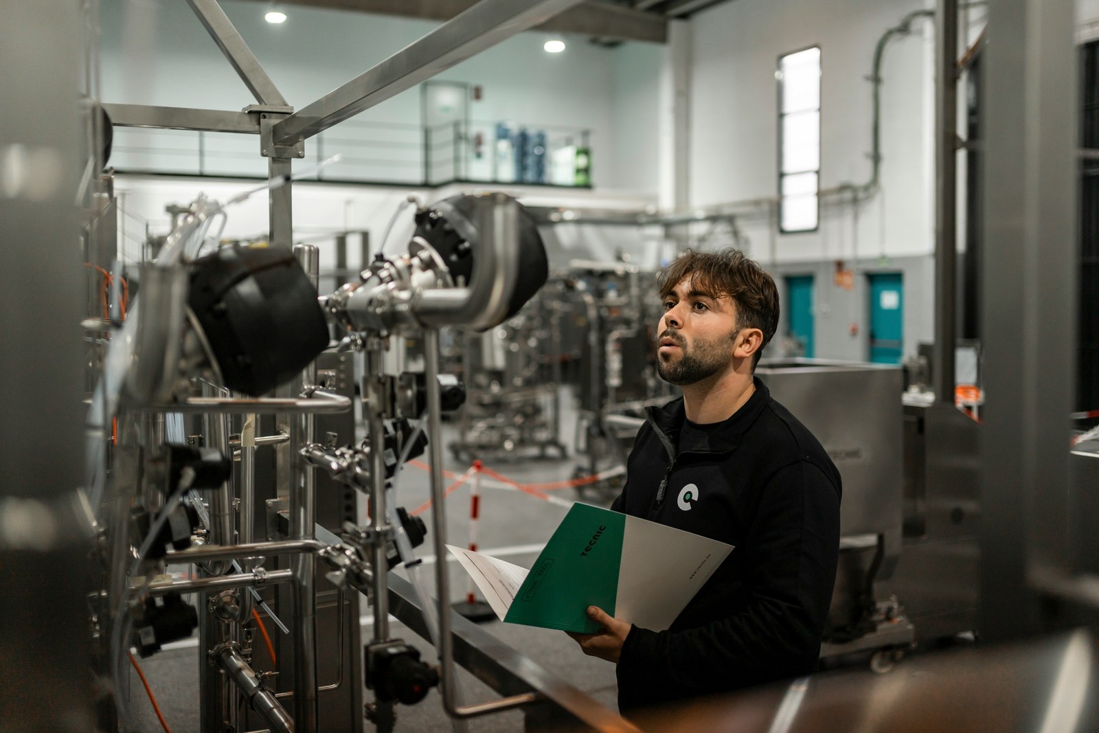 An engineer on a plant floor, looking up from the documentation in his hands to the stainless steel pipework in front of him